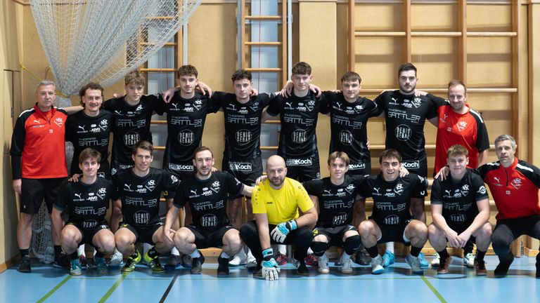 Eine Gruppe junger Männer in schwarzen Trikots mit Sponsorenlogo posiert für ein Mannschaftsfoto auf einem Spielfeld. Der Trainer im gelben Shirt kniet in der Mitte.