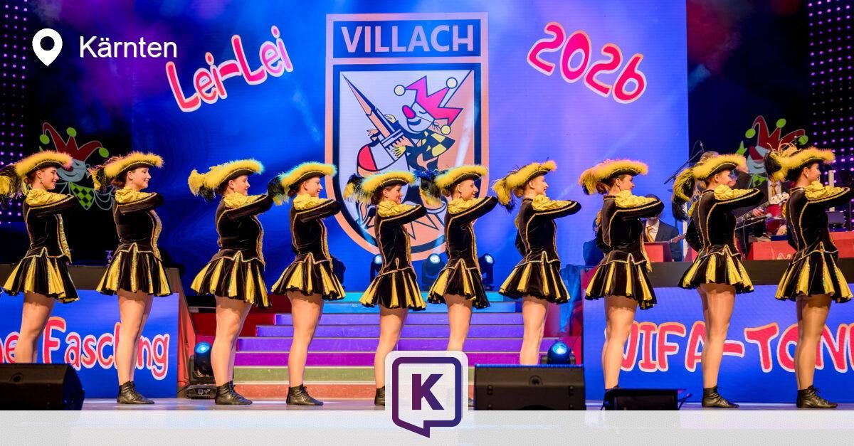 Eine Gruppe von Künstlern in schwarzen und gelben Kostümen mit breitkrempigen Hüten und Stiefeln tritt auf einer Bühne auf, vor einem großen 'Villach 2026'-Banner und mit Lichtern im Hintergrund.