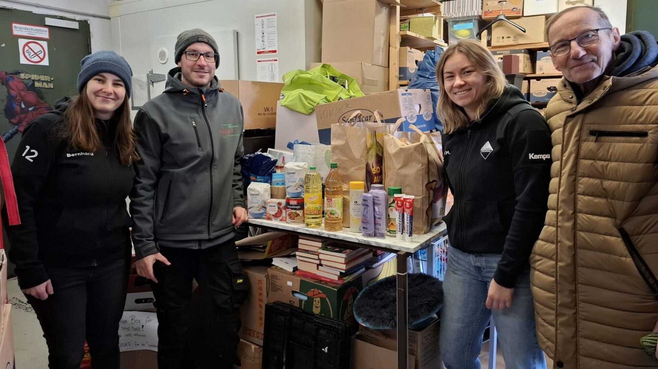Zwei Personen stehen lächelnd in einem Raum mit Kisten. Ein Tisch mit Büchern und Lebensmitteln steht zwischen ihnen. Die Person links trägt eine Jacke, und die Person rechts trägt eine Kapuzenjacke.