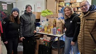 Zwei Personen stehen lächelnd in einem Raum mit Kisten. Ein Tisch mit Büchern und Lebensmitteln steht zwischen ihnen. Die Person links trägt eine Jacke, und die Person rechts trägt eine Kapuzenjacke.