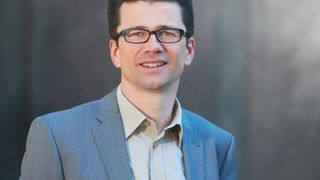 Ein Mann in einem blauen Anzug und mit Brille lächelt in die Kamera. Er steht vor einer hellgrauen Wand.