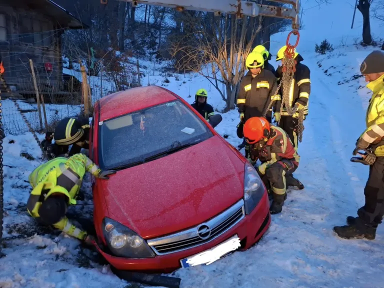 Feuerwehrleute in Sicherheitsausrüstung verwenden Ketten, um ein rotes Auto aus dem Schnee zu ziehen. Das Fahrzeug hat ein Nummernschild.