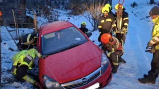 Feuerwehrleute in Sicherheitsausrüstung verwenden Ketten, um ein rotes Auto aus dem Schnee zu ziehen. Das Fahrzeug hat ein Nummernschild.