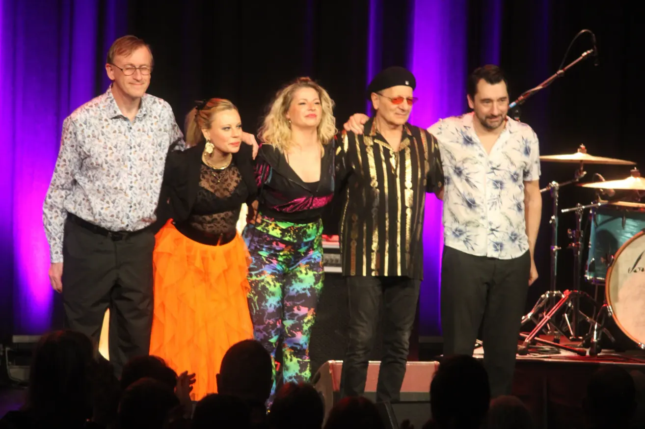 Fünf Personen stehen auf einer Bühne, zwei Frauen in farbenfrohen Outfits, eine in Schwarz, zwei Männer, einer mit Hut und Sonnenbrille, ein Mikrofon auf einem Ständer im Hintergrund und eine Menge vor ihnen.
