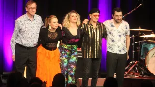 Fünf Personen stehen auf einer Bühne, zwei Frauen in farbenfrohen Outfits, eine in Schwarz, zwei Männer, einer mit Hut und Sonnenbrille, ein Mikrofon auf einem Ständer im Hintergrund und eine Menge vor ihnen.