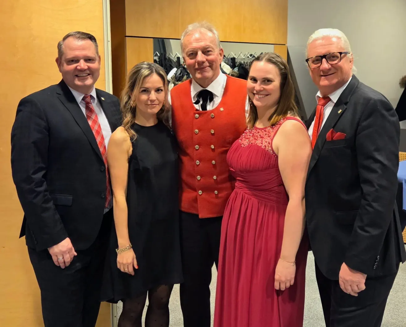 Fünf Personen stehen für ein Foto zusammen, alle in formeller Kleidung. Zwei Männer und drei Frauen lächeln in die Kamera. Der Mann in der Mitte trägt eine rote Weste mit Knöpfen. Hinter ihnen ist eine Holzwand und ein Spiegel mit Kleidern zu sehen.