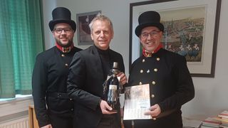 Drei Männer in Uniform, einer hält eine Flasche und ein Papier, ein anderer hält eine Puppe und lächelt, und der dritte lächelt und schaut in die Kamera. Hinter ihnen ist eine Wand mit Bilderrahmen.