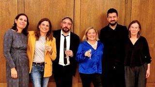 Vier Personen posieren für ein Foto vor einem hölzernen Hintergrund. Sie alle lächeln. Die Frau auf der linken Seite trägt eine gelbe Jacke und hält ein Weinglas. Der Mann in der Mitte trägt einen Anzug und eine Krawatte. Die Frau auf der rechten Seite trägt eine blaue Bluse und hält ein Weinglas. Der Mann ganz rechts trägt ein schwarzes Shirt.