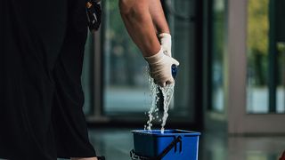 Eine Person reinigt einen Boden mit einem Mopp, trägt Handschuhe und hält einen blauen Eimer. Wasser spritzt aus dem Eimer.