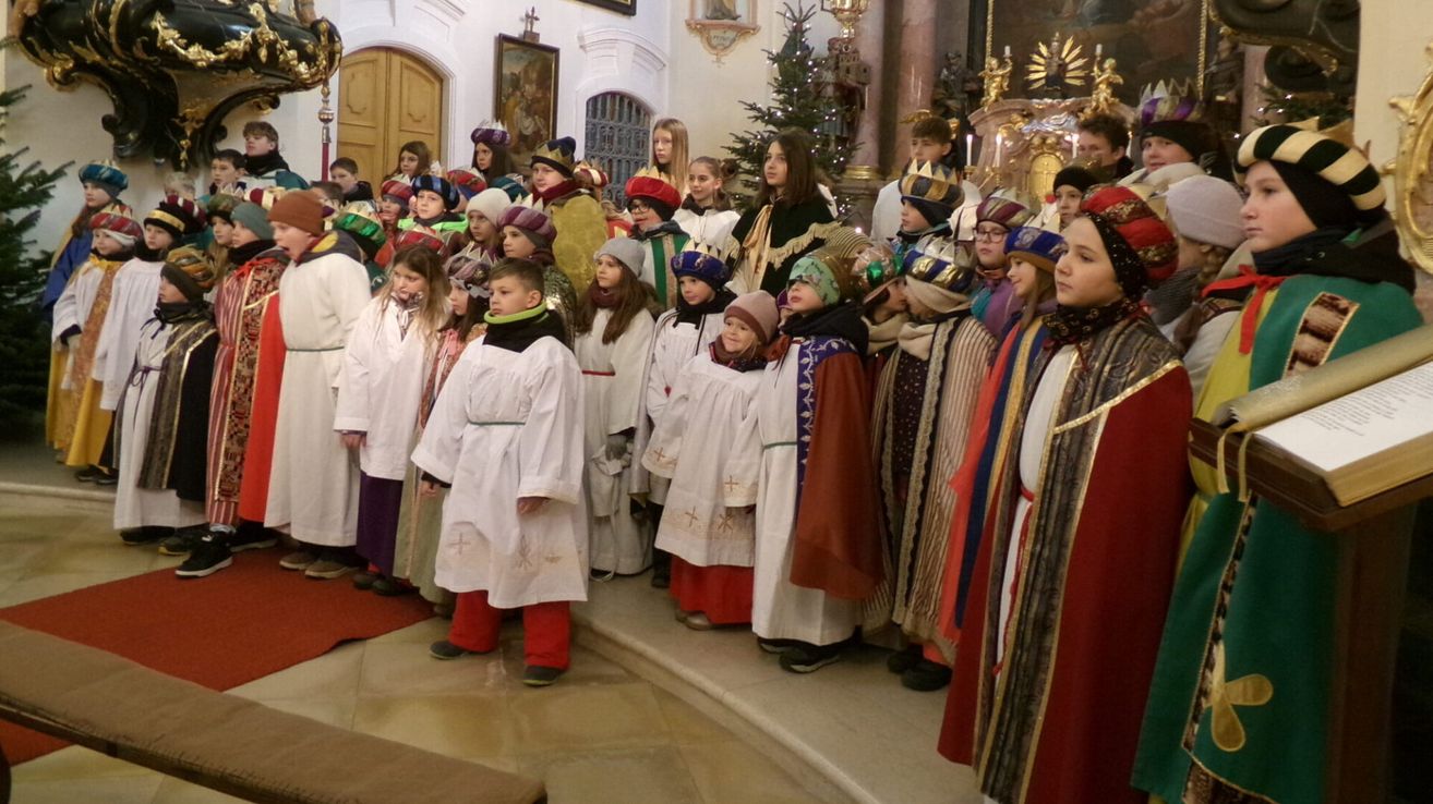 Eine Gruppe von Kindern in verschiedenen Kostümen und Mützen steht in einer Kirche, möglicherweise für eine Aufführung oder Feier.