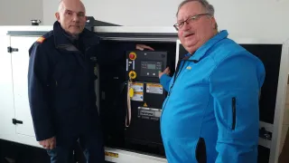 Zwei Männer in Jacken stehen neben einer Maschine, einer mit Brille und der andere mit einer Plakette an seiner Jacke.