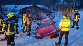 Ein rotes Auto steckt im Schnee fest, mit einem Feuerwehrmann in der Nähe. Das Auto ist teilweise im Schnee vergraben. Hinter dem Auto befindet sich ein Holzhaus und ein Zaun.
