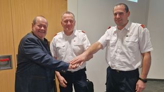 Drei Männer, zwei in Uniform, lächeln und schütteln sich die Hände in einem Raum mit einer weißen Wand.