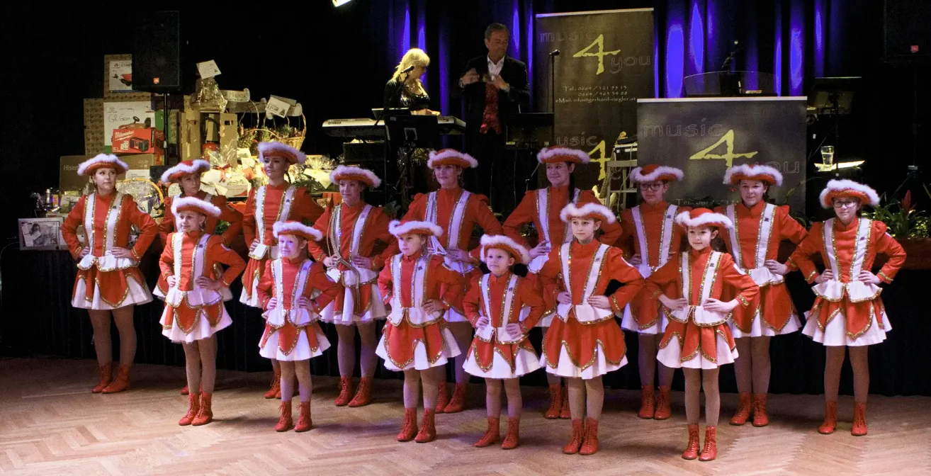 Eine Gruppe junger Kinder in roten und weißen Kostümen mit Hüten tritt auf der Bühne auf. Dahinter stehen ein Mann und eine Frau mit einem Klavier und einem Mikrofon.