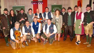 Eine Gruppe von Musikern posiert für ein Foto auf einer Bühne und hält verschiedene Instrumente wie Gitarre, Trompete und Saxophon. Sie sind in traditioneller deutscher Kleidung gekleidet, und einige tragen Hüte.