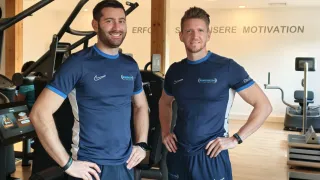 Zwei Männer stehen in einem Fitnessstudio und tragen blaue Trikots mit Nike-Logos. Sie lächeln und scheinen für ein Foto zu posieren. Hinter ihnen ist eine weiße Wand mit Text, und verschiedene Fitnessgeräte sind sichtbar.