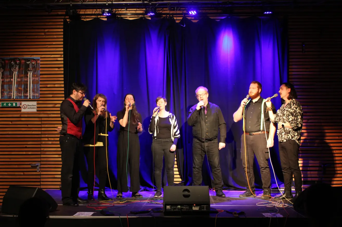 Eine Gruppe von Sängern steht mit Mikrofonen und Musikequipment auf der Bühne. Sie tragen alle schwarze Kleidung. Die Bühne hat einen blauen Vorhang als Hintergrund mit lila Beleuchtung.