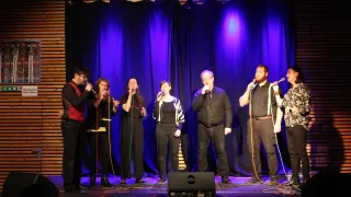 Eine Gruppe von Sängern steht mit Mikrofonen und Musikequipment auf der Bühne. Sie tragen alle schwarze Kleidung. Die Bühne hat einen blauen Vorhang als Hintergrund mit lila Beleuchtung.