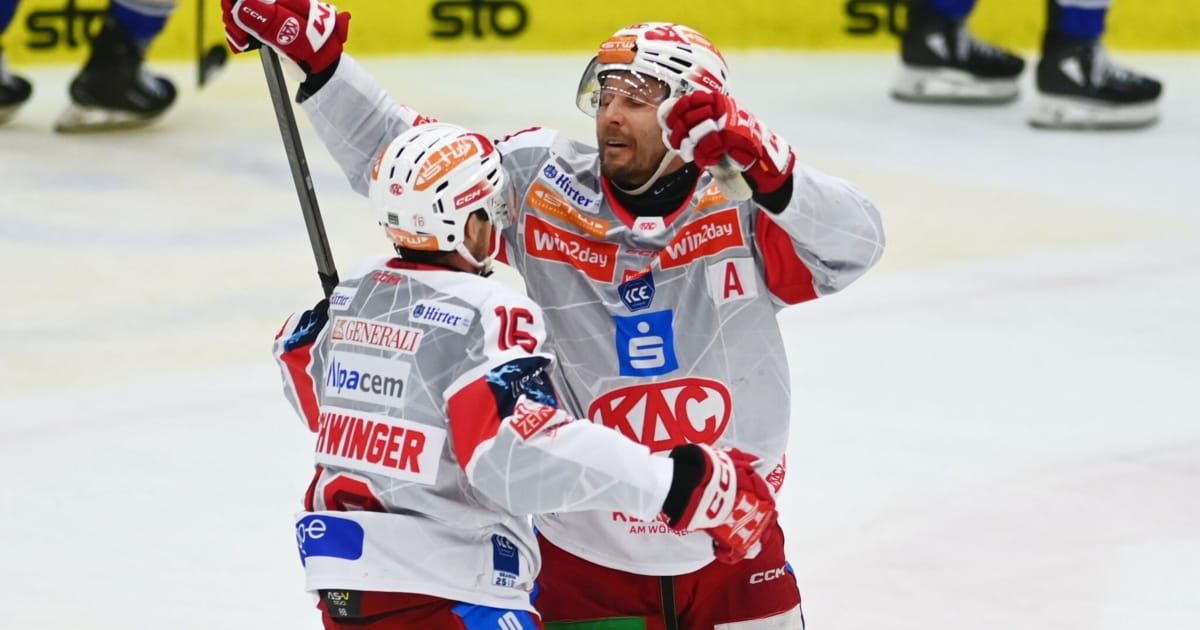 Zwei Eishockeyspieler feiern auf dem Eis, einer hält einen Hockeyschläger und der andere hat beide Hände erhoben. Sie tragen rote und weiße Trikots mit verschiedenen Logos.