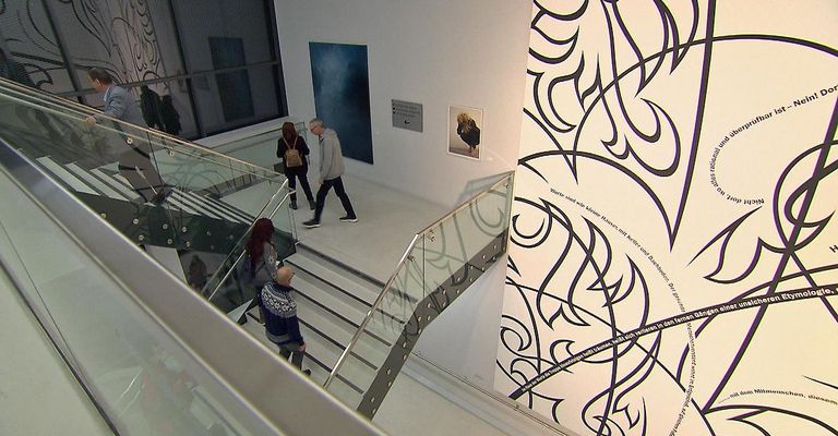 Im Inneren einer Kunstgalerie gehen mehrere Personen auf einer Glastreppe. An der Wand befindet sich ein großes Kunstwerk mit schwarzem und weißem Muster.