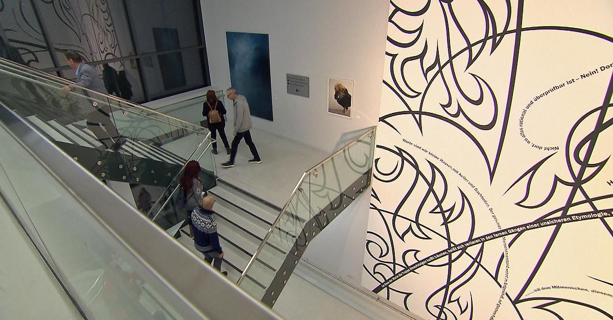 Im Inneren einer Kunstgalerie gehen mehrere Personen auf einer Glastreppe. An der Wand befindet sich ein großes Kunstwerk mit schwarzem und weißem Muster.