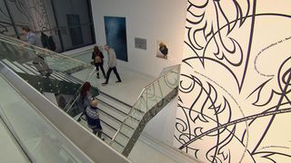 Im Inneren einer Kunstgalerie gehen mehrere Personen auf einer Glastreppe. An der Wand befindet sich ein großes Kunstwerk mit schwarzem und weißem Muster.
