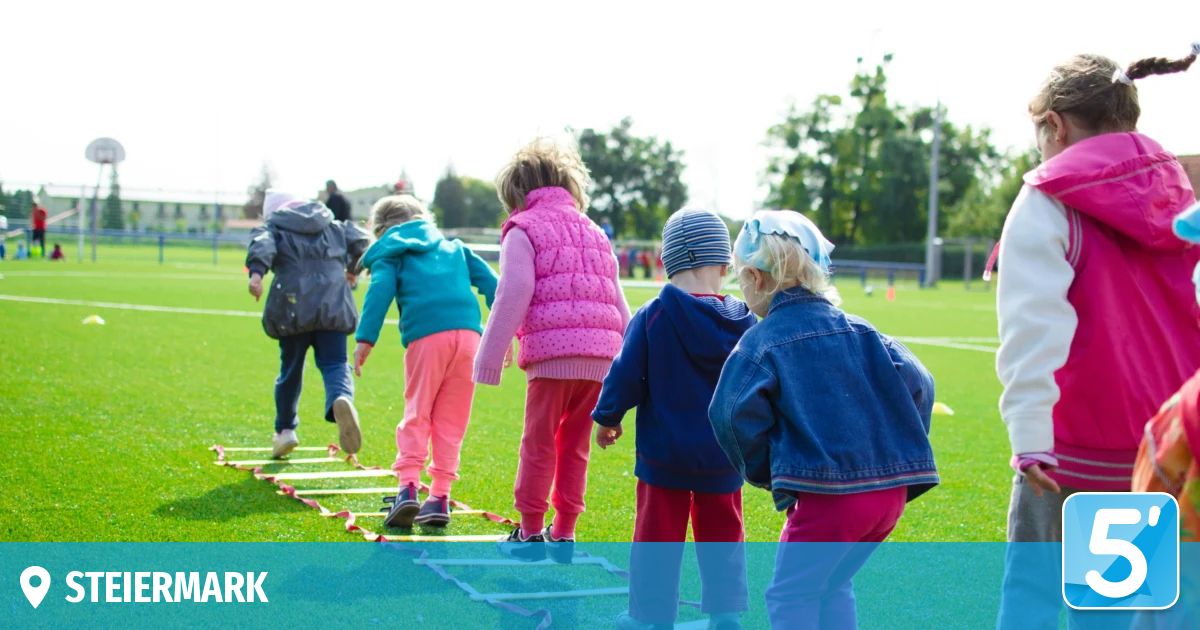 Eine Gruppe von Kindern übt auf einem Grasfeld mit Hürden Geschicklichkeitsübungen. Einige tragen Jacken und Turnschuhe.