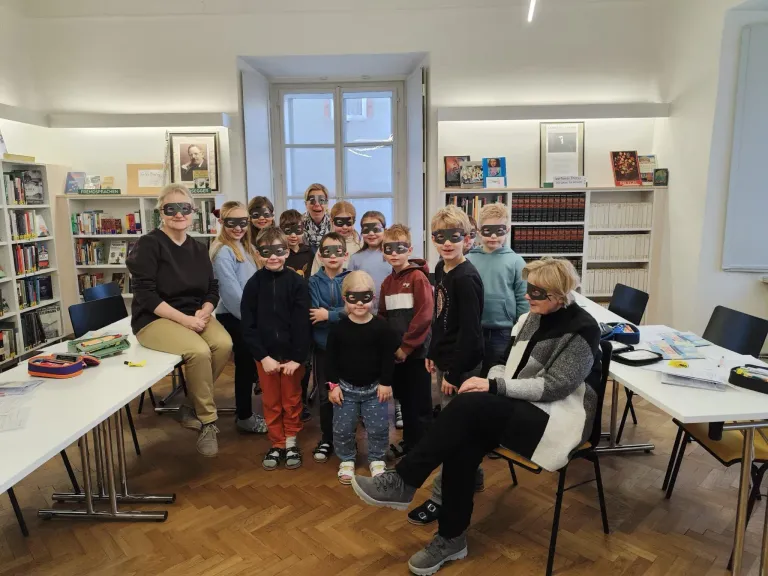 Eine Gruppe von Kindern mit verbundenen Augen posiert für ein Foto in einer Bibliothek. Zwei Erwachsene sitzen auf beiden Seiten. Im Hintergrund sind Bücherregale und Tische zu sehen.