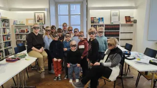 Eine Gruppe von Kindern mit verbundenen Augen posiert für ein Foto in einer Bibliothek. Zwei Erwachsene sitzen auf beiden Seiten. Im Hintergrund sind Bücherregale und Tische zu sehen.