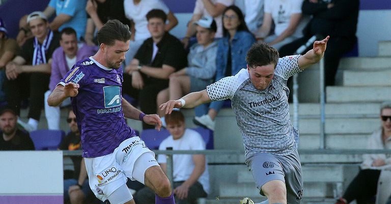 Zwei Fußballspieler streiten um einen Ball. Einer trägt ein lila Trikot, der andere grau. Sie sind von Zuschauern umgeben, die auf Tribünen sitzen.