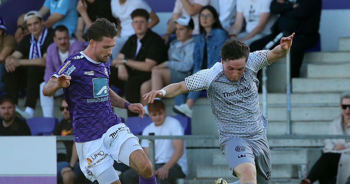 Zwei Fußballspieler streiten um einen Ball. Einer trägt ein lila Trikot, der andere grau. Sie sind von Zuschauern umgeben, die auf Tribünen sitzen.