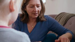 Eine Frau mit braunen Haaren sitzt auf einer Couch und scheint mit jemandem zu sprechen. Sie trägt ein blaues Oberteil und eine Halskette mit einem Herzanhänger.