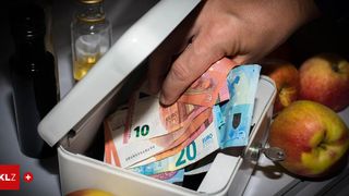 Eine Hand nimmt einen 20-Euro-Schein aus einer Box, die mit verschiedenen Euro-Scheinen und einer Flasche Öl gefüllt ist.