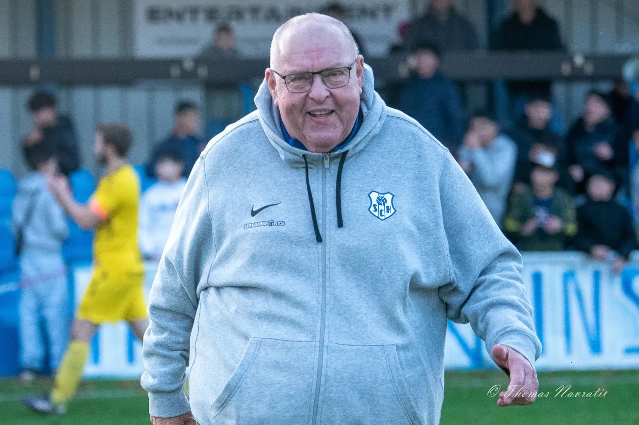 Ein älterer Mann mit Brille und grauer Kapuzenjacke läuft auf einem Fußballfeld. Hinter ihm sehen mehrere Personen ein Spiel zu.