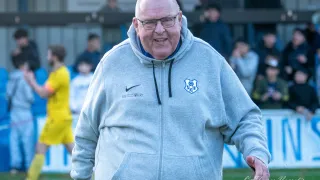 Ein älterer Mann mit Brille und grauer Kapuzenjacke läuft auf einem Fußballfeld. Hinter ihm sehen mehrere Personen ein Spiel zu.