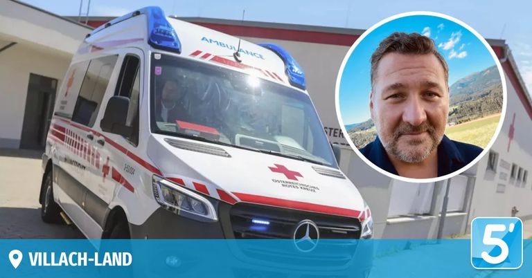 Ein Krankenwagen mit dem Wort 'Ambulanz' auf dem Dach und einem roten Kreuzsymbol an der Seite, mit einer Person im Inneren und einem Mann in einem blauen Hemd neben ihm.