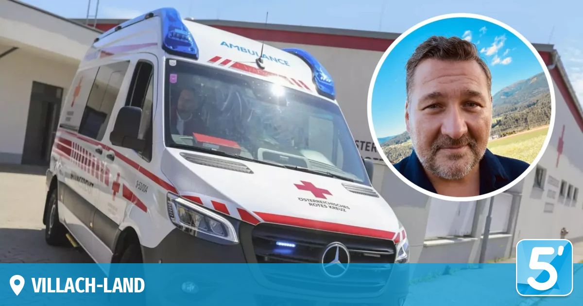 Ein Krankenwagen mit dem Wort 'Ambulanz' auf dem Dach und einem roten Kreuzsymbol an der Seite, mit einer Person im Inneren und einem Mann in einem blauen Hemd neben ihm.
