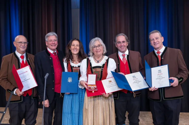 Sechs Menschen in traditioneller Kleidung stehen gemeinsam auf einer Bühne und lächeln für ein Foto. Sie halten verschiedene Urkunden in den Händen. Eine Frau in der Mitte trägt eine Halskette. Ein Mikrofon und Vorhänge sind im Hintergrund zu sehen.