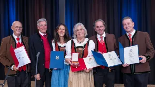 Sechs Menschen in traditioneller Kleidung stehen gemeinsam auf einer Bühne und lächeln für ein Foto. Sie halten verschiedene Urkunden in den Händen. Eine Frau in der Mitte trägt eine Halskette. Ein Mikrofon und Vorhänge sind im Hintergrund zu sehen.