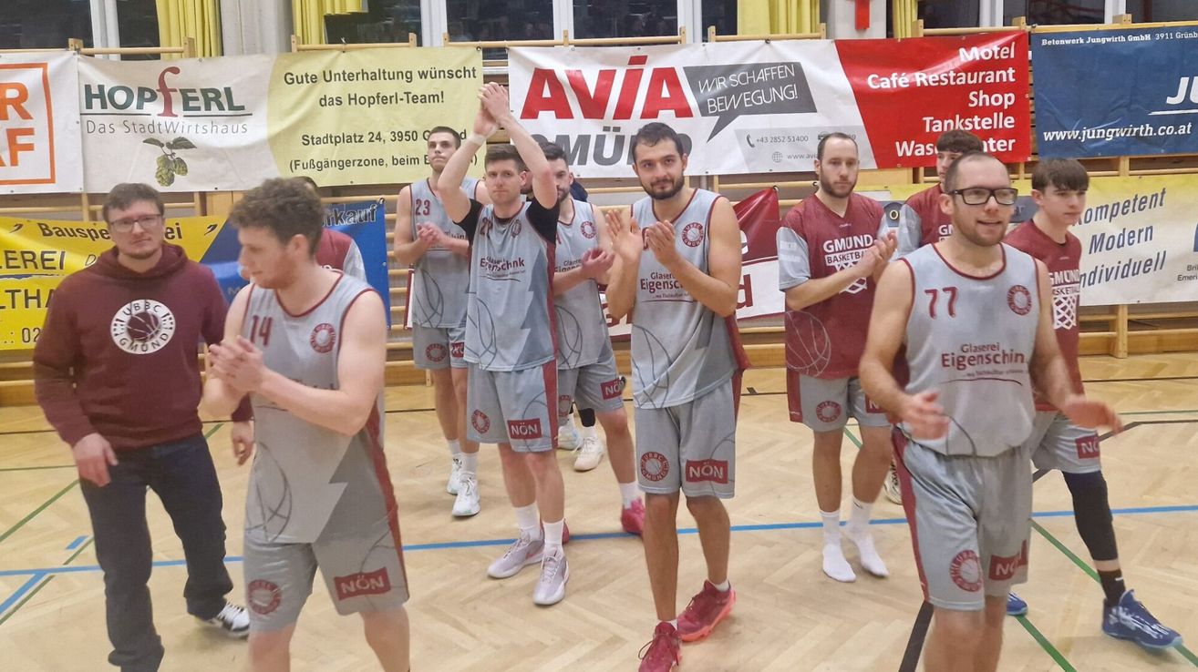 Eine Basketballmannschaft steht auf einem Spielfeld. Sie tragen graue Uniformen mit roten Besätzen. Ein Mann hebt die Hände, während ein anderer klatscht. Ein Banner mit der Aufschrift 'AVIA' ist im Hintergrund sichtbar.