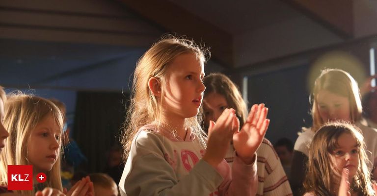 Ein junges Mädchen mit blonden Haaren klatscht in die Hände und schaut etwas an. Hinter ihr steht ein weiteres Mädchen, das ebenfalls klatscht. Sie sind beide drinnen.