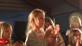 Ein junges Mädchen mit blonden Haaren klatscht in die Hände und schaut etwas an. Hinter ihr steht ein weiteres Mädchen, das ebenfalls klatscht. Sie sind beide drinnen.