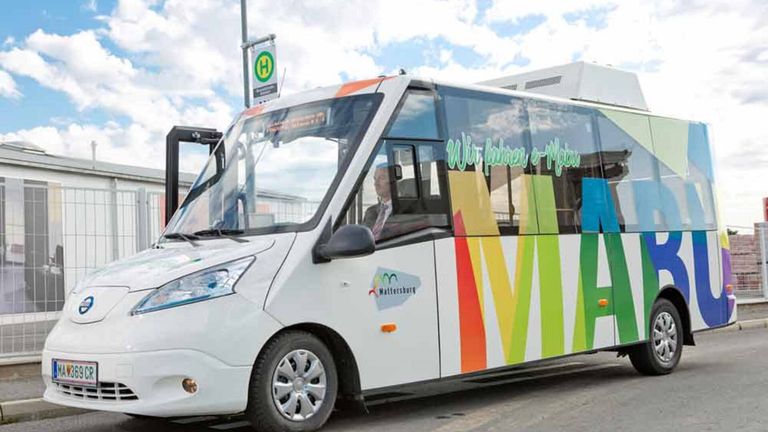 Ein weißer Bus mit einem bunten Design an der Seite steht mit der Aufschrift 'Wir fahren e-Mobil'. Ein Mann sitzt auf dem Fahrersitz, und der Bus ist am Straßenrand geparkt.