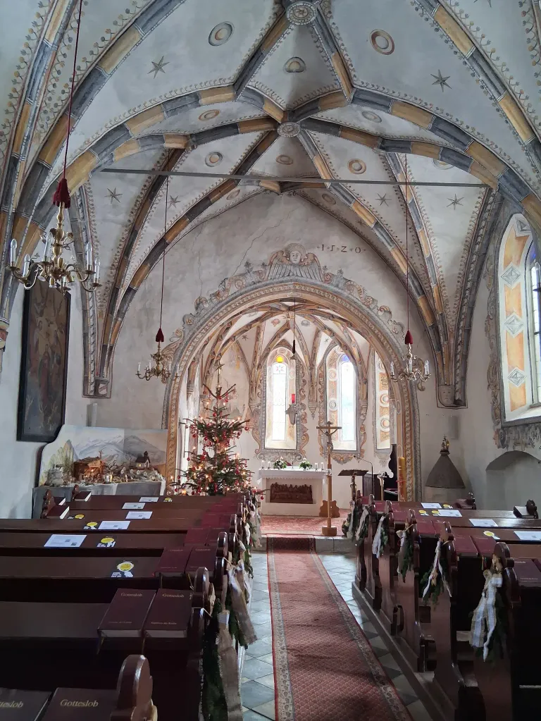 Ein Innenansicht einer Kirche mit Weihnachtsbaum, Kerzen, einem Kreuz, einem roten Teppich und hölzernen Kirchenbänken. Die Decke ist mit aufwendigen Mustern verziert.