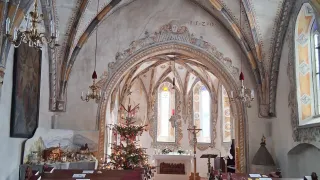 Ein Innenansicht einer Kirche mit Weihnachtsbaum, Kerzen, einem Kreuz, einem roten Teppich und hölzernen Kirchenbänken. Die Decke ist mit aufwendigen Mustern verziert.