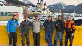 Eine Gruppe von sechs Männern in Winterkleidung steht lächelnd vor einer gelben Wand, um ein Foto zu machen. Hinter ihnen ist ein Van mit dem Wort 'INKU' zu sehen. Im Hintergrund bilden Gebäude und verschneite Berge eine malerische Winterlandschaft.