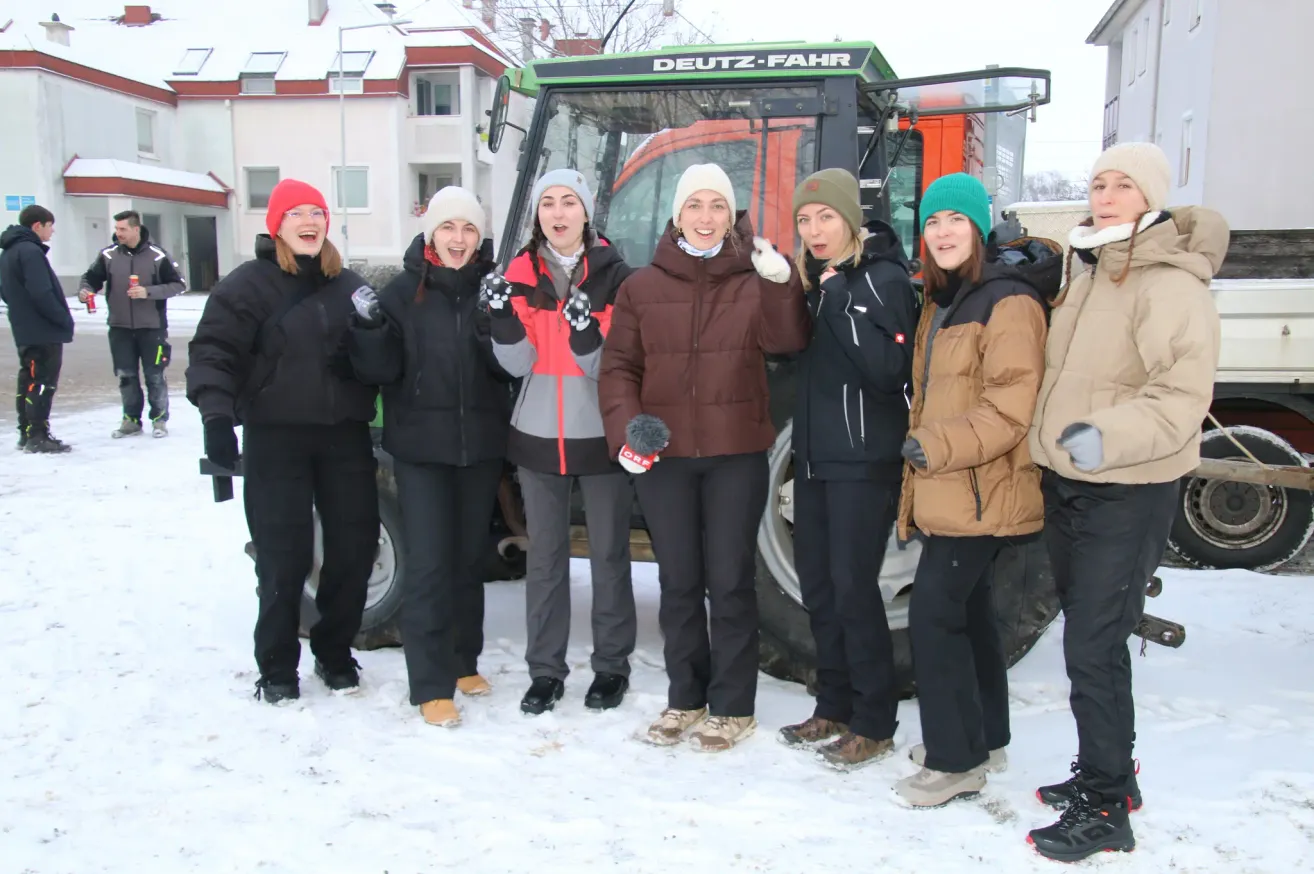Eine Gruppe von Frauen posiert für ein Foto im Schnee vor einem Traktor mit der Aufschrift 'DEUTZ-FAHR'. Sie sind alle warm in Winterkleidung gekleidet.