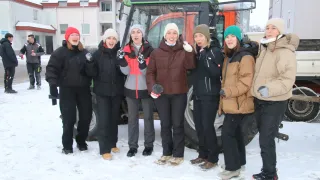 Eine Gruppe von Frauen posiert für ein Foto im Schnee vor einem Traktor mit der Aufschrift 'DEUTZ-FAHR'. Sie sind alle warm in Winterkleidung gekleidet.