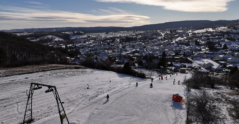 Luftaufnahme einer verschneiten Piste mit mehreren Skifahrern. Gebäude im Hintergrund und ein bewölkter Himmel.