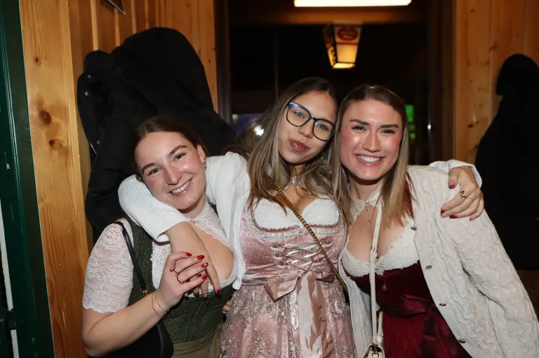 Drei Frauen lächeln und posieren für ein Foto. Die Frau auf der linken Seite trägt einen grünen Pullover und eine schwarze Tasche. Die Frau in der Mitte trägt einen weißen Pullover und eine Brille. Die Frau auf der rechten Seite trägt ein rotes Kleid.